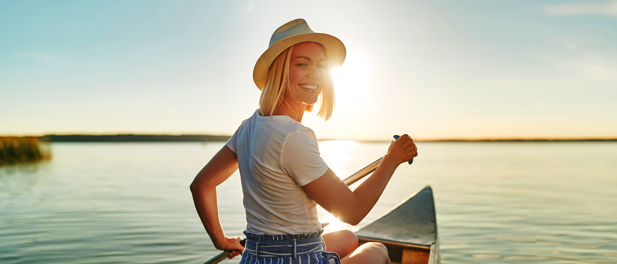 Lächelnde Frau mit Strohhut beim Rudern in einem Kanu, genießt die Sonne auf einem ruhigen Gewässer während des Sonnenuntergangs.