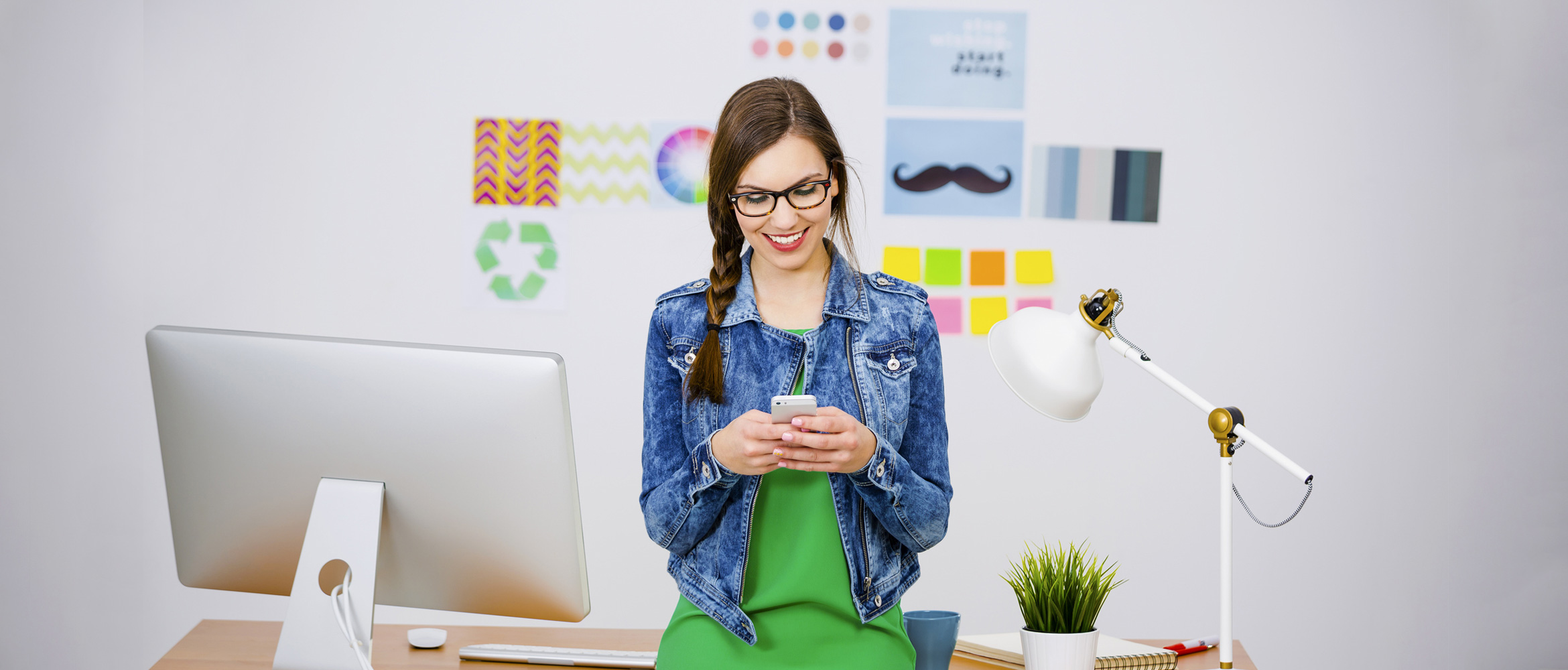 Eine junge Frau mit Brille und einem lächelnden Gesichtsausdruck steht vor einem Schreibtisch mit einem iMac, einer weißen Schreibtischlampe und diversen Büroutensilien. Sie trägt ein grünes Top und eine blaue Jeansjacke und hält ein Smartphone in ihren Händen. Im Hintergrund sind bunte Wanddekorationen und Notizzettel zu sehen.