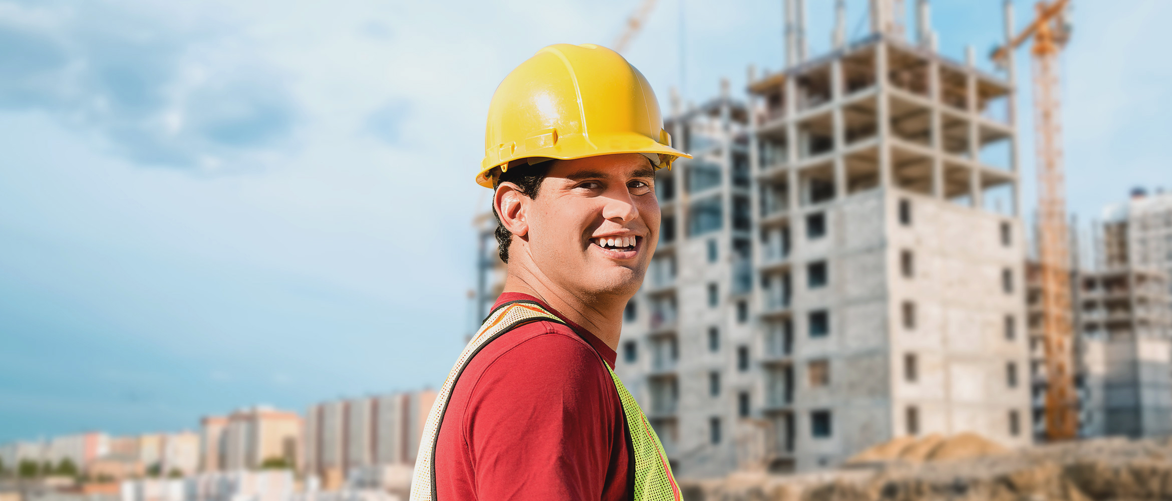 Ein lächelnder Bauarbeiter mit gelbem Helm und reflektierender Weste steht vor einer Baustelle, das Bild porträtiert Arbeitszufriedenheit und Sicherheitsbewusstsein im Baugewerbe.