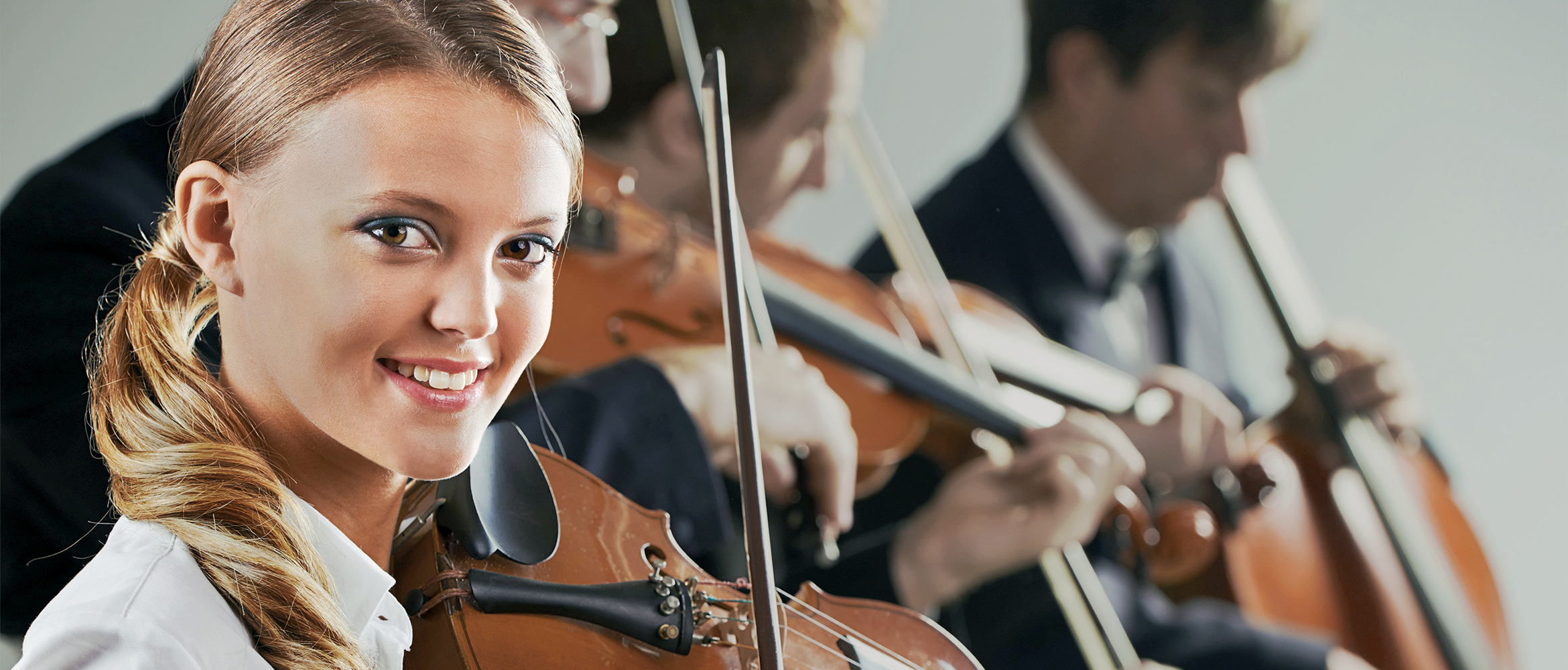 Eine junge Frau mit zurückgebundenen Haaren spielt lächelnd eine Violine, im Hintergrund verschwommen weitere Geiger, was eine musikalische Ensemble-Performance suggeriert.