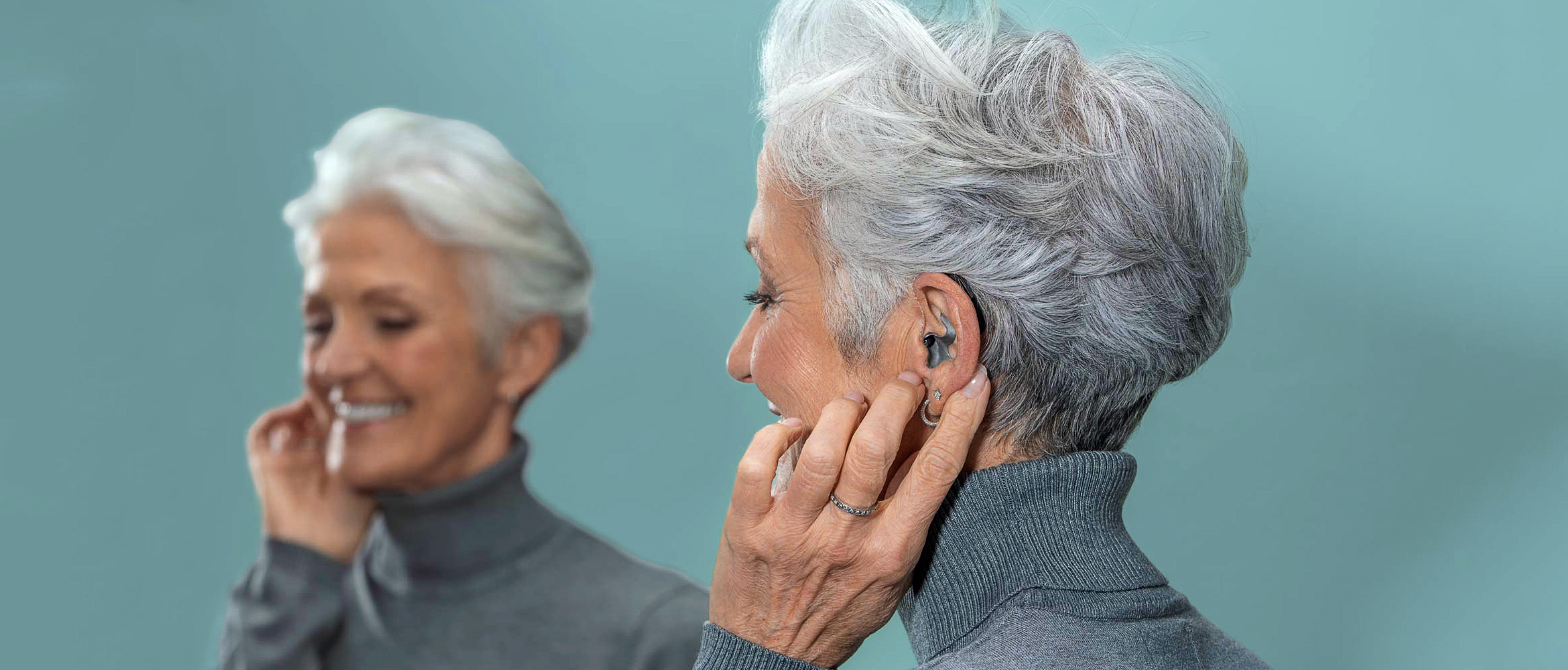 Ein lächelnde ältere Frau mit silbergrauem Haar setzt ein modernes, titanfarbenes Hörgerät ein.