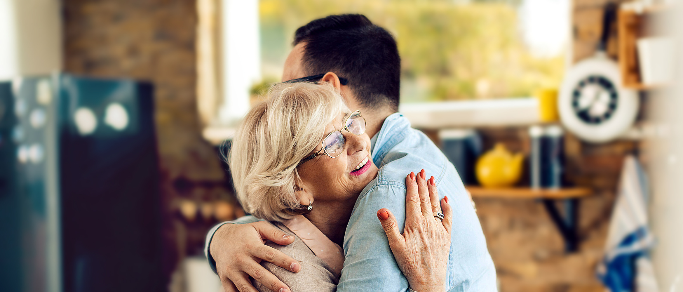 Eine ältere Dame mit grauen Haaren und Brille umarmt herzlich einen jüngeren Mann in einem gemütlichen Wohnzimmer, wobei ein Gefühl von familiärer Zuneigung und Wärme vermittelt wird.