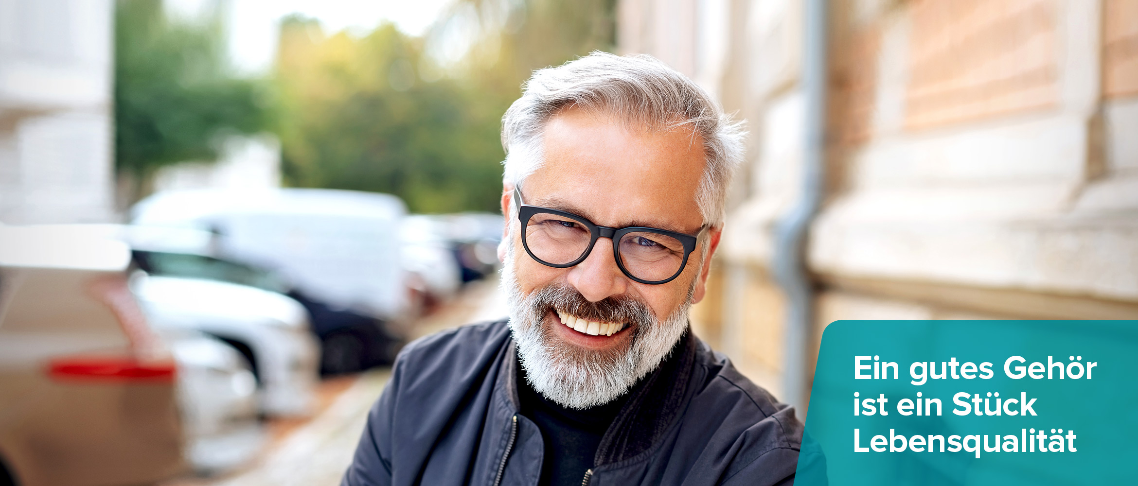 Ein fröhlicher Mann mit grauem Bart und Brille lächelt in die Kamera, auf der rechten Seite steht 'Ein gutes Gehör ist ein Stück Lebensqualität' über einem türkisen Banner.