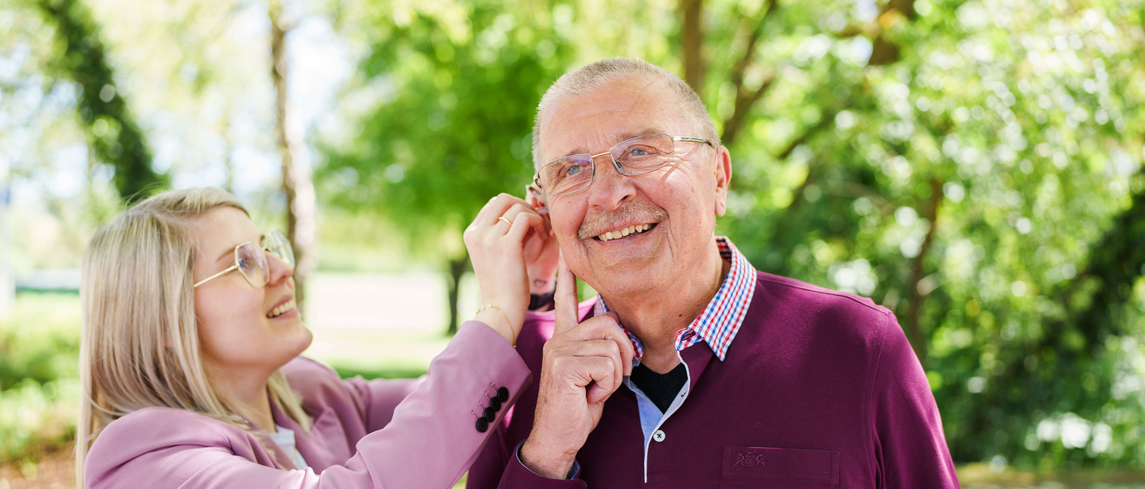 Ein älterer Herr mit Brille und einem Lächeln wird von einer jüngeren Frau mit blonden Haaren und Brille beim Anpassen eines Hörgeräts unterstützt. Sie befinden sich in einem parkähnlichen Umfeld mit Bäumen im Hintergrund.
