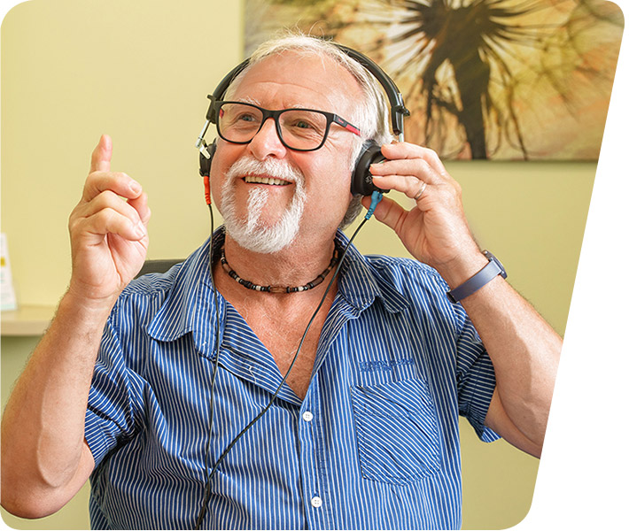 Fröhlicher älterer Mann mit grauem Bart, der ein Headset trägt und den Daumen nach oben zeigt, was Zufriedenheit und gute Hörqualität dank Picard Hörgeräten symbolisiert. Er trägt ein blau gestreiftes Hemd und sitzt vor einem unscharfen Hintergrund mit einer Wanddekoration.